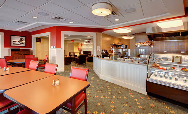 Interior view of a dining area in a senior living facility with wooden tables and red chairs. To the right is a food service counter with a glass display case containing desserts and other food items. The room has a patterned carpet and a ceiling with recessed lighting. In the background, there is a lounge area with armchairs and framed artwork on the walls.