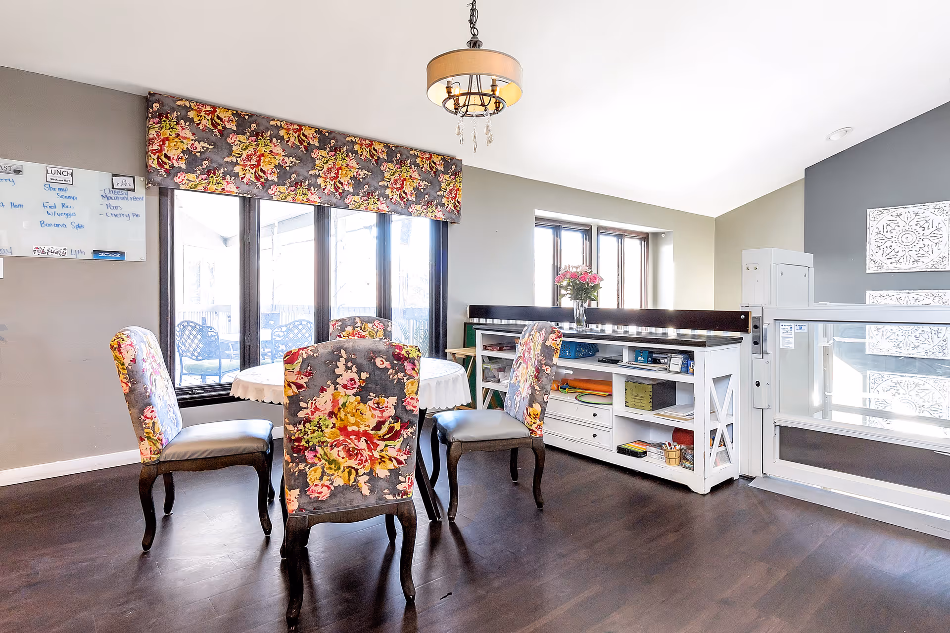 Bright dining area with a round table and four floral-upholstered chairs by large windows and a white storage cabinet.