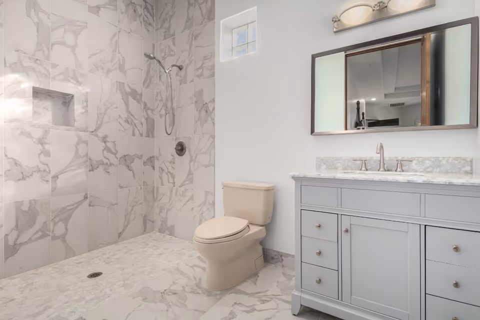 Modern bathroom with marble-tiled walk-in shower, a beige toilet, and a gray vanity with mirror and sink.