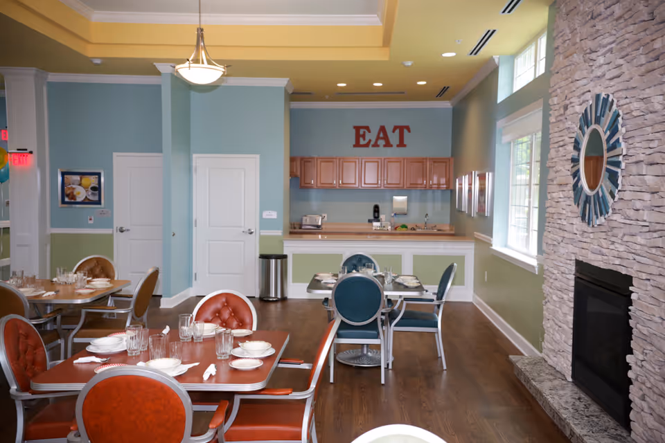 A dining room with several tables set with plates, bowls, glasses, and napkins. The room features a stone fireplace with a decorative round mirror above it, large windows letting in natural light, and a kitchen area in the background with wooden cabinets and the word 'EAT' displayed on the wall.