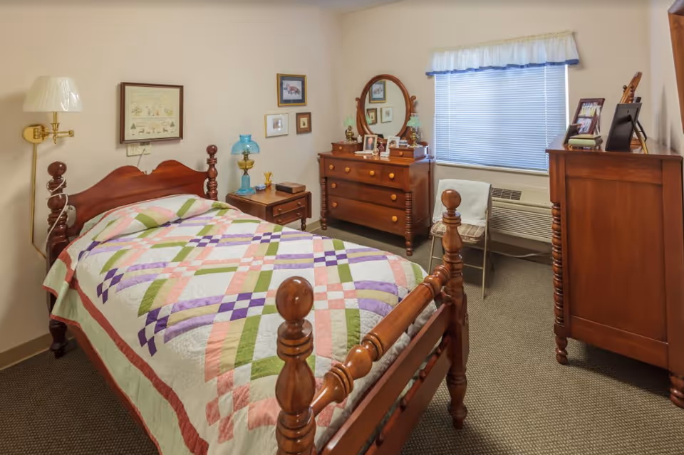 A cozy bedroom with a wooden bed frame covered by a colorful patchwork quilt. The room features a wooden dresser with a round mirror, a small side table with a blue lamp, and a wooden chest of drawers. There is a window with blinds and a white valance, and a folding chair with a cushion is placed near the window. The walls have framed pictures and the floor is carpeted.