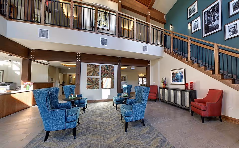 A spacious and well-lit senior living facility common area with high ceilings and a second-floor balcony. The room features four blue upholstered armchairs arranged around small tables on a patterned carpet, two red armchairs near a sideboard with decorative items, and framed artwork on the walls. A staircase with wooden railings leads to the upper floor.