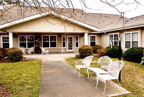 Outdoor patio area of a senior living facility with white metal chairs and tables on a concrete walkway. The building has beige siding, multiple windows, and a covered porch. There are bushes and grass surrounding the walkway, and leafless tree branches are visible overhead.