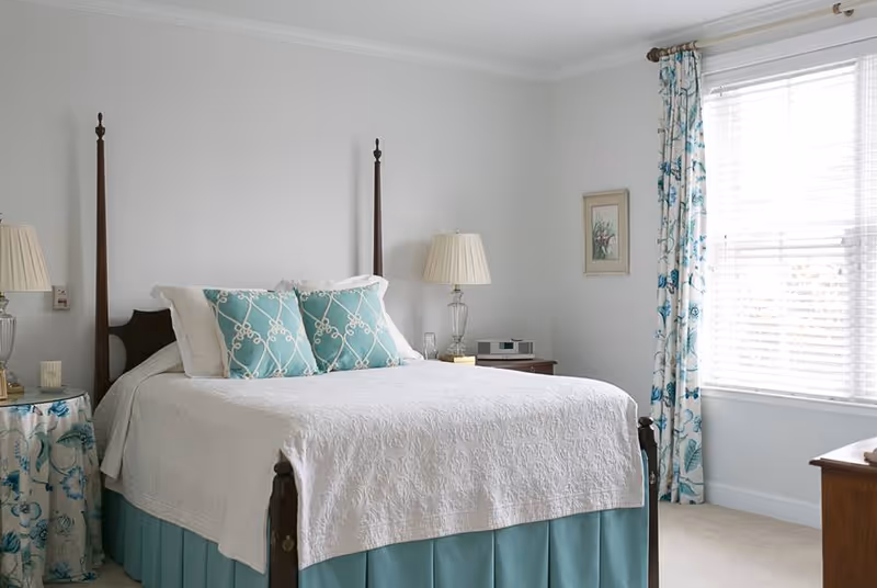 A neatly made bed with a white quilt and two decorative blue pillows in a bright bedroom. The bed has a dark wooden frame with tall posts. There are two bedside tables with lamps, one round table covered with a floral cloth and the other a wooden table with a clock radio. A window with white blinds and blue floral curtains lets in natural light. A small framed picture hangs on the wall.