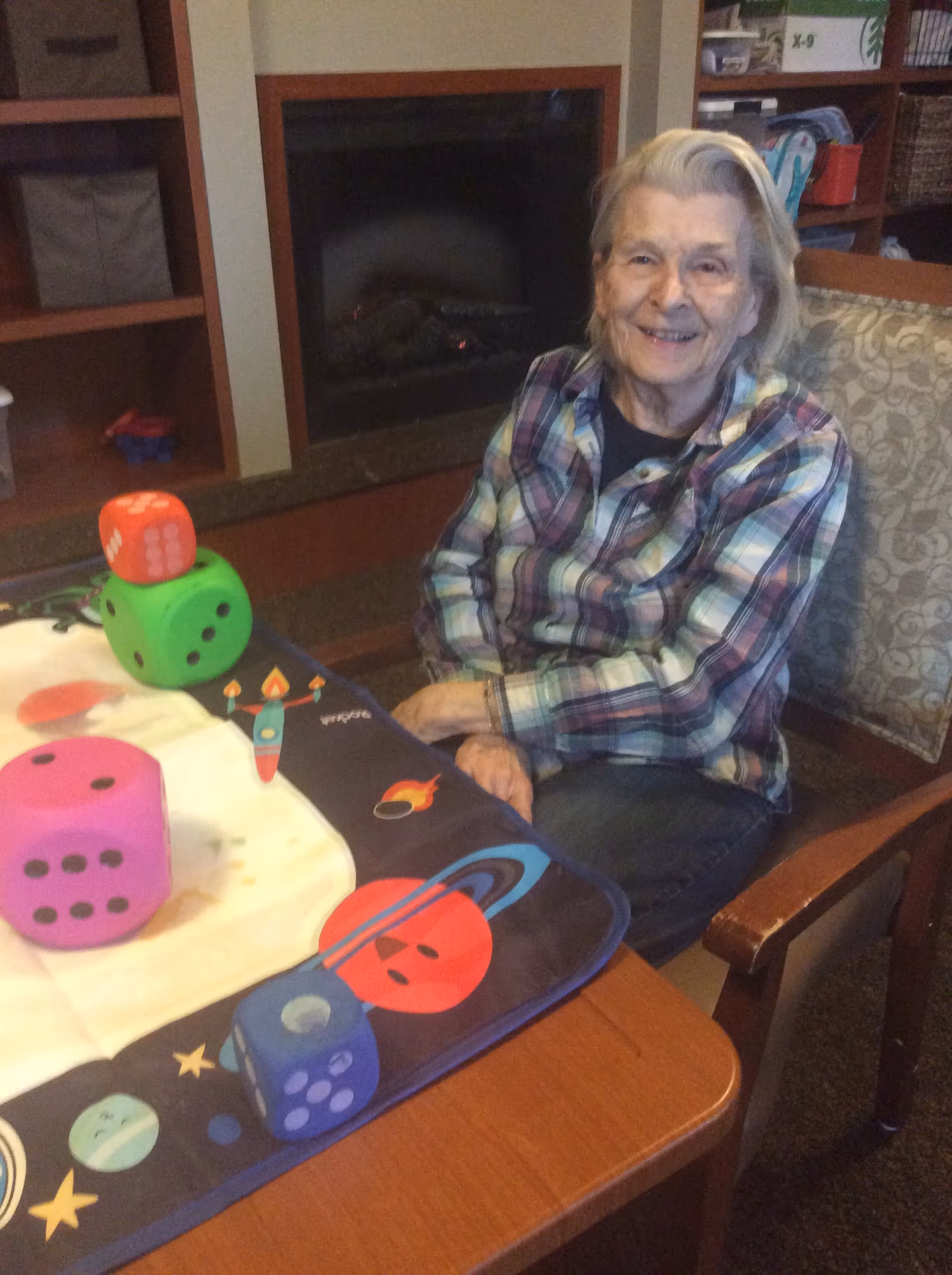 An elderly woman with gray hair wearing a plaid shirt sits at a table with a colorful space-themed tablecloth. On the table are large foam dice in pink, green, red, and blue. Behind her is a fireplace and shelving with various items.