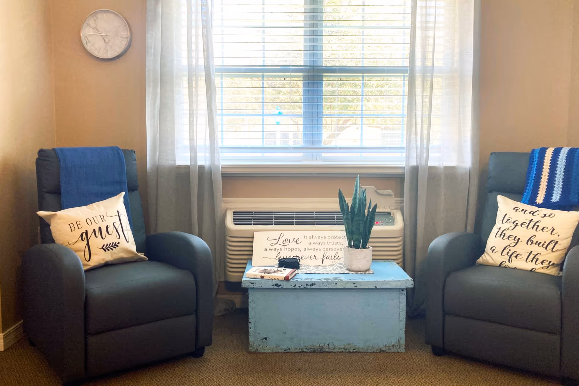 A cozy sitting area with two dark gray armchairs, each with a decorative pillow and a blue throw blanket. Between the chairs is a distressed blue wooden chest serving as a table, holding a potted plant, a small sign with an inspirational quote, and some books. Behind the chest is a window with sheer curtains and a wall-mounted air conditioning unit below it. A round wall clock is visible on the left wall.