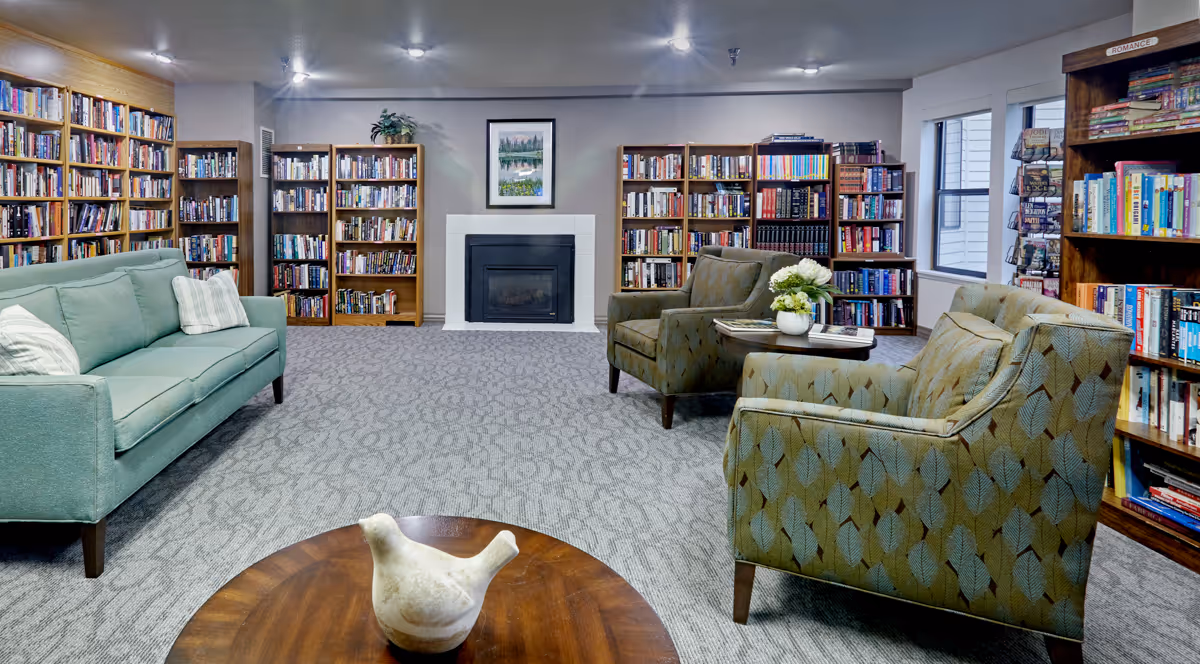 A cozy senior living facility library with multiple wooden bookshelves filled with books along the walls. There is a light green sofa on the left, two patterned armchairs on the right, and a wooden coffee table with a decorative bird sculpture in the foreground. A fireplace with a framed picture above it is centered on the back wall. The room is carpeted and well-lit with ceiling lights.