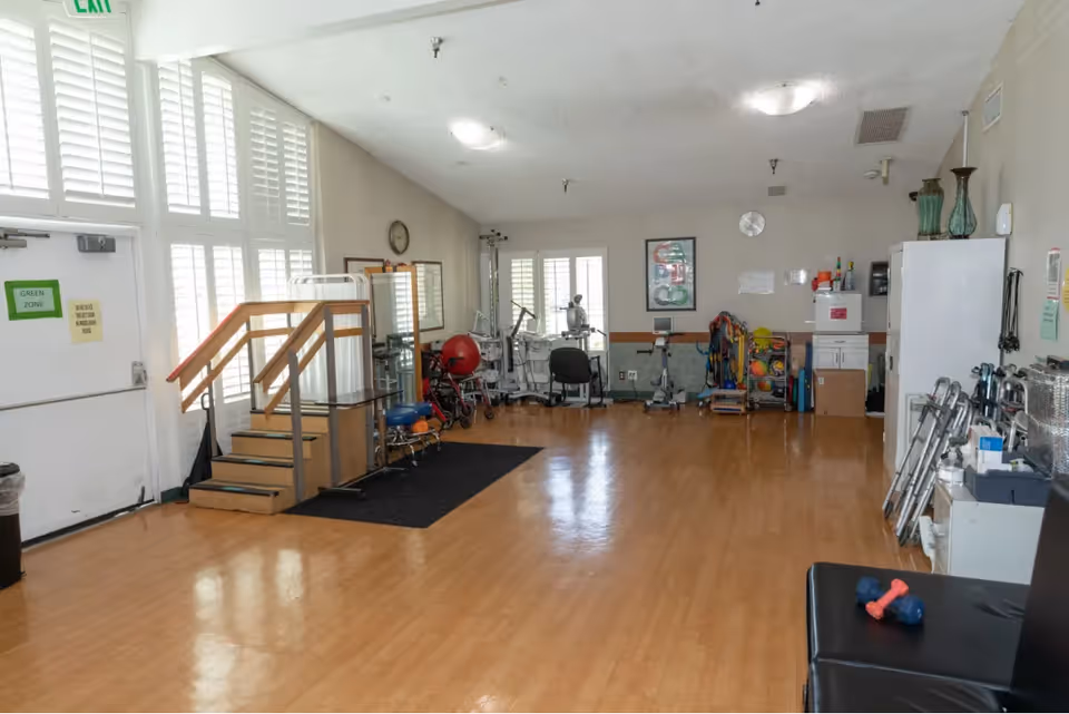 A spacious room with wooden flooring and large windows with white shutters allowing natural light. The room contains various physical therapy and exercise equipment including parallel bars, exercise balls, walkers, and weights. There is a small set of stairs with handrails on the left side near the exit door marked with a green zone sign. The walls are light-colored with a clock and framed pictures, and the ceiling has multiple light fixtures.