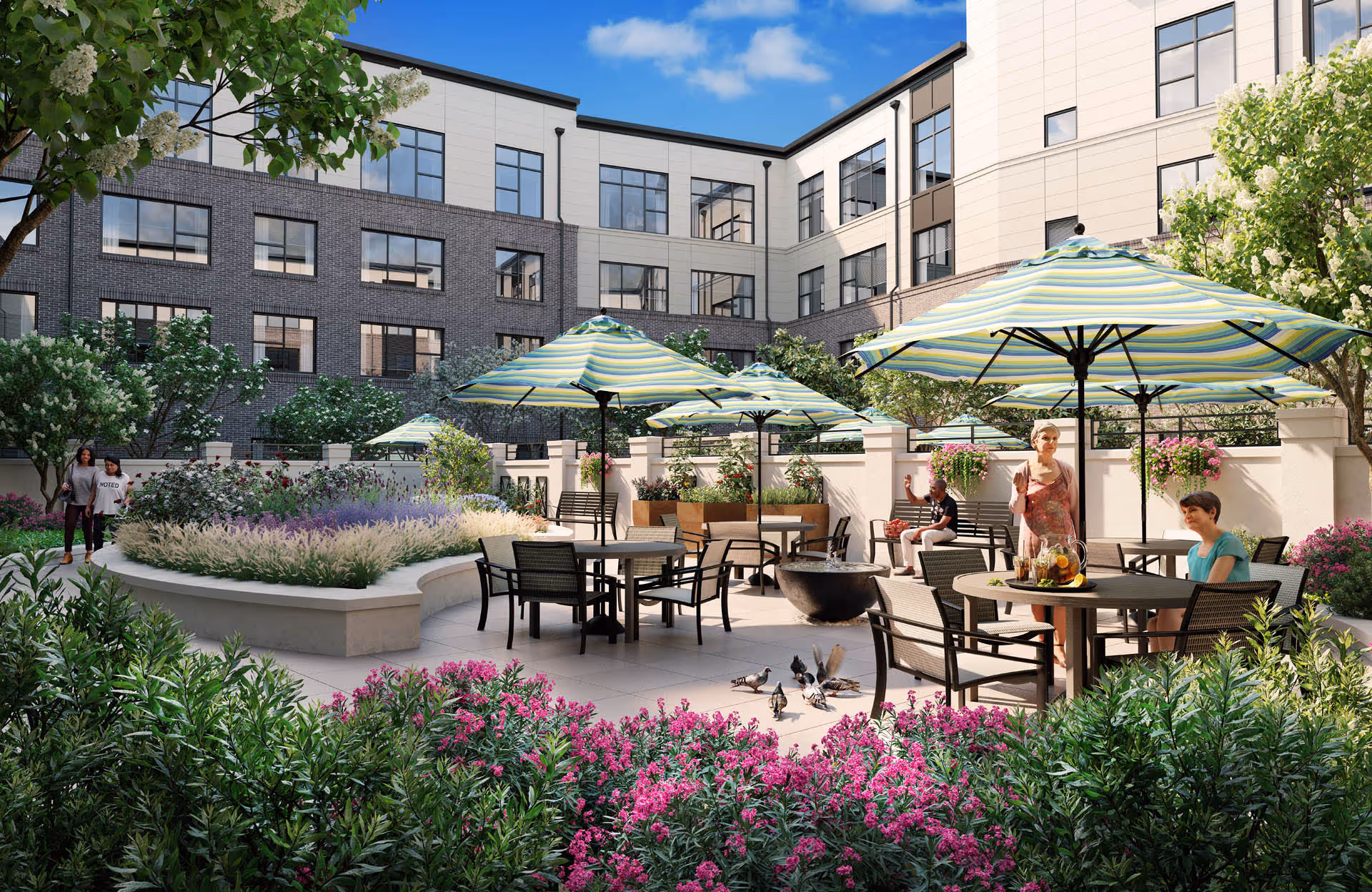 Outdoor courtyard area of a senior living facility with several tables and chairs under large striped umbrellas. People are sitting and standing around the tables, surrounded by lush greenery, flowering plants, and trees. The building with multiple windows forms the backdrop under a blue sky with some clouds.