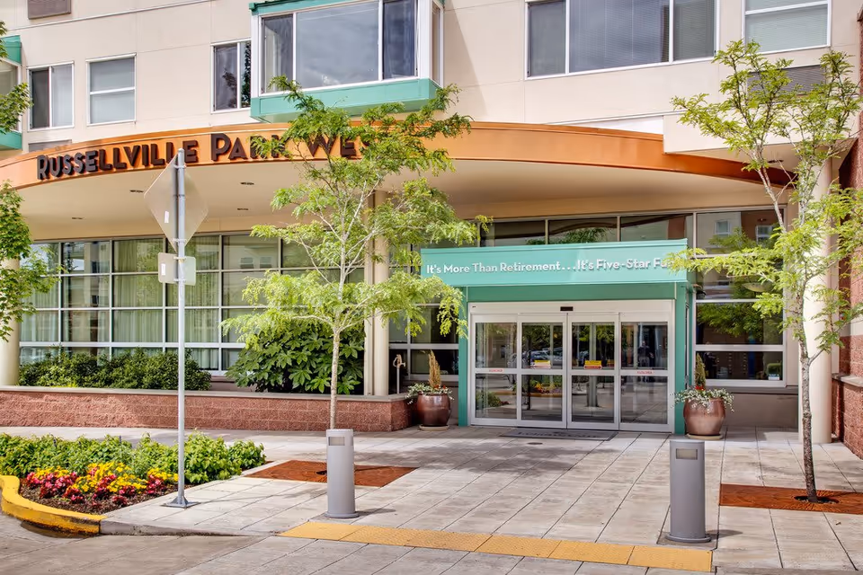Entrance to Russellville Park facility with automatic sliding glass doors, a green awning with the text 'It's More Than Retirement... It's Five-Star Fu', trees, and landscaped flower beds in front.