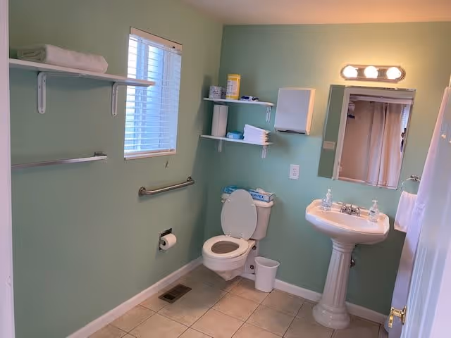 A clean bathroom with light green walls, a white toilet with the lid open, a white pedestal sink with two soap dispensers, a mirror with three lights above it, a window with white blinds, and shelves holding towels and cleaning supplies.