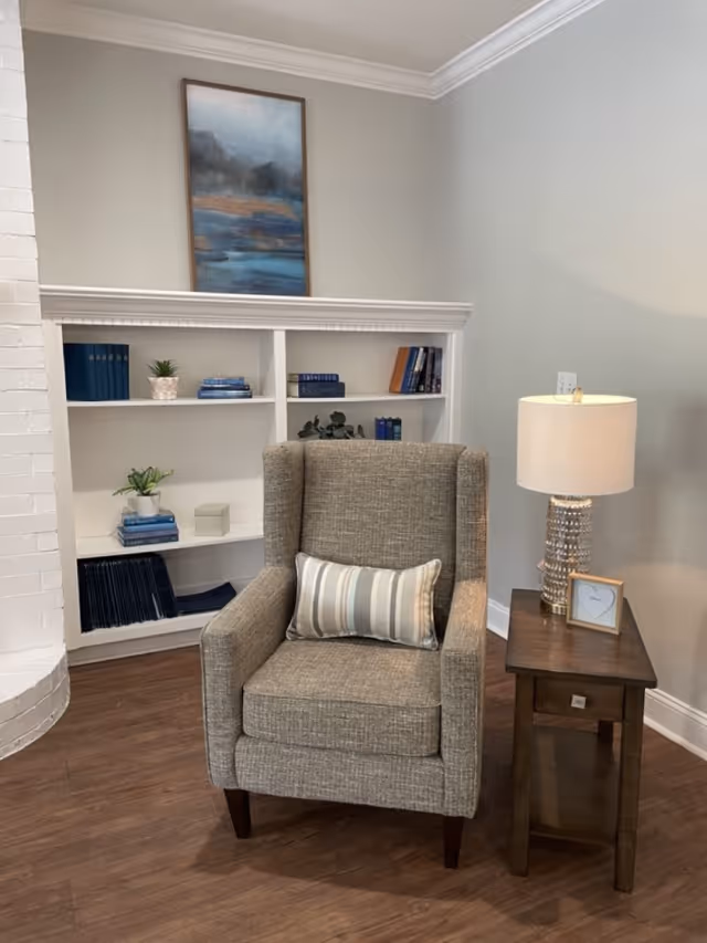 A cozy corner of a living room featuring a gray upholstered armchair with a striped lumbar pillow, a wooden side table with a decorative lamp and a small framed picture, and a white built-in bookshelf with books and small plants. A framed abstract painting hangs above the bookshelf, and the floor is wood with light gray walls.