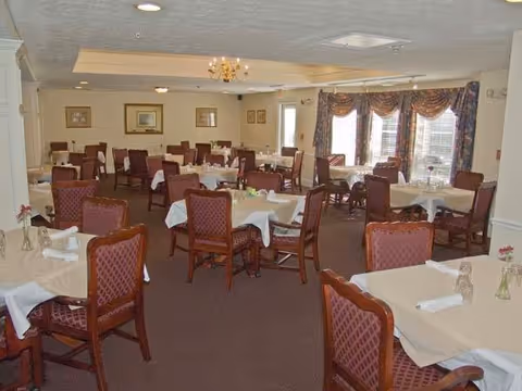 Formal dining room with multiple tables draped in tablecloths and upholstered chairs arranged near windows.