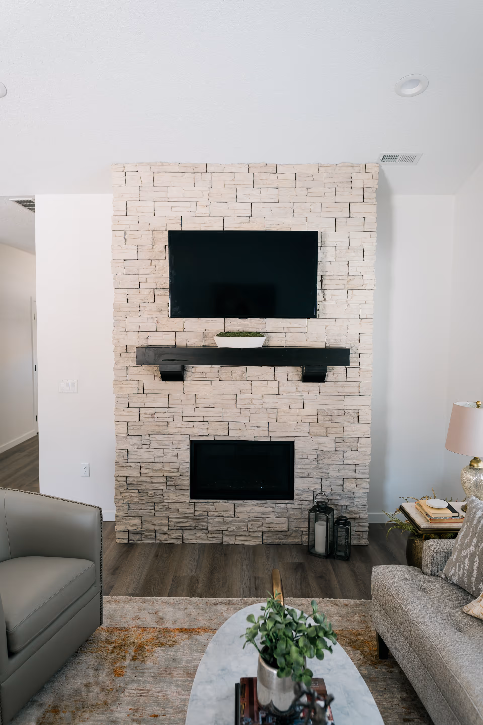 A cozy living room featuring a stone fireplace with a mounted flat-screen TV above it. Below the TV is a black mantel with a small decorative plant. To the right of the fireplace are two lanterns on the floor. The room has a gray armchair on the left, a gray sofa on the right with a side table holding a lamp and books, and a marble coffee table with a potted plant in the foreground. The floor is wood with a patterned area rug.