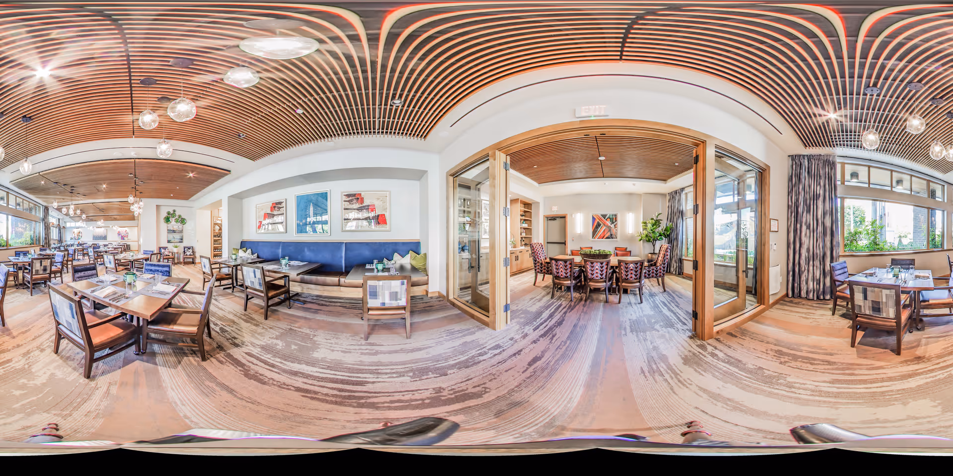 A spacious and well-lit dining area with wooden tables and chairs, featuring a modern wooden slatted ceiling with hanging light fixtures. The room has large windows allowing natural light to fill the space. There is a separate private dining room visible through glass doors, furnished with a large round table and chairs. The decor includes framed artwork on the walls and plants, creating a welcoming atmosphere.