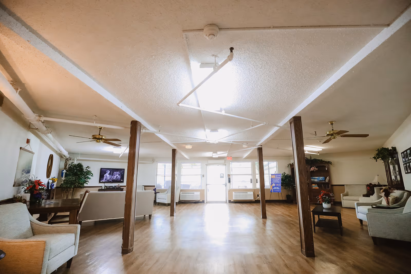 A spacious common area in a senior living facility with wooden floors, several armchairs and sofas arranged around coffee tables, ceiling fans, potted plants, and a television mounted on the wall. The room is well-lit with natural light coming through large windows and a glass door at the far end.