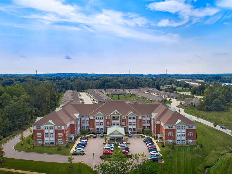 Aerial view of Summit Corners senior living facility showing a large, three-story brick building with a peaked roof and multiple windows. The building is surrounded by a parking lot with cars and landscaped green areas. In the background, there are additional smaller buildings and a wooded area under a partly cloudy blue sky.