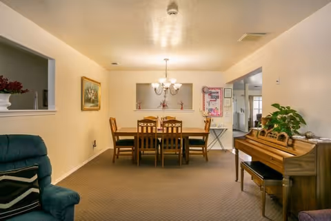 A dining room with a wooden dining table and six chairs under a chandelier light fixture. To the right, there is a wooden piano with a plant on top. The walls are light-colored, and there is a framed painting on the left wall. A green upholstered chair with a patterned pillow is partially visible on the left side. There is a pass-through window in the wall behind the dining table, and an open doorway leading to another room on the right.