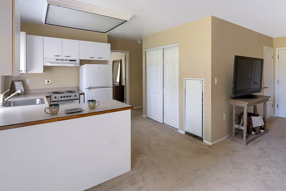 Interior view of a senior living facility apartment showing a small kitchen area with white cabinets, a stove, and a refrigerator. There is a counter with two coffee mugs and a small tray. Adjacent to the kitchen is a living area with a TV on a wooden stand, beige carpet, and beige walls. A closet with white sliding doors is visible in the background.