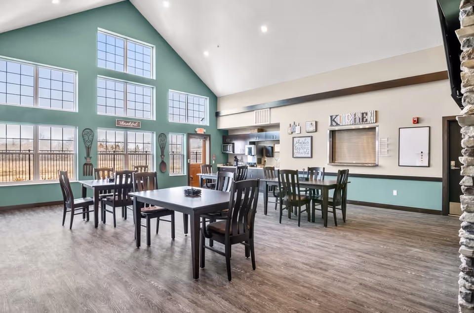 A bright and spacious dining area with multiple dark wooden tables and chairs. Large windows with a green accent wall allow natural light to fill the room. The room features a high vaulted ceiling with recessed lighting. On one wall, there is a serving window labeled 'KITCHEN' and decorative signs including one that says 'Gather Grateful Hearts'. The floor is covered with wood-like flooring, and there is a stone pillar partially visible on the right side.