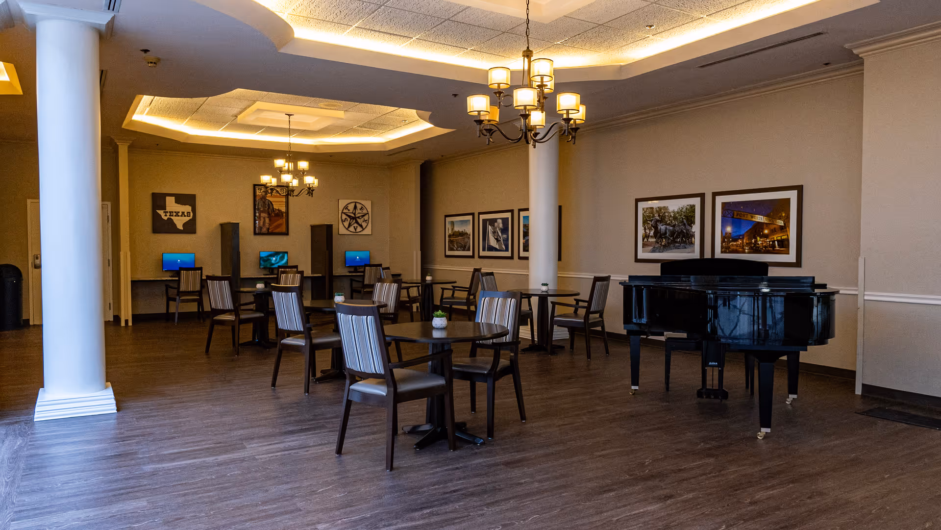 A spacious common area with wooden flooring, several tables and chairs arranged for seating, a black grand piano on the right side, and a row of computer stations with chairs along the back wall. The room is decorated with framed pictures on the walls and has warm ceiling lighting with chandeliers.