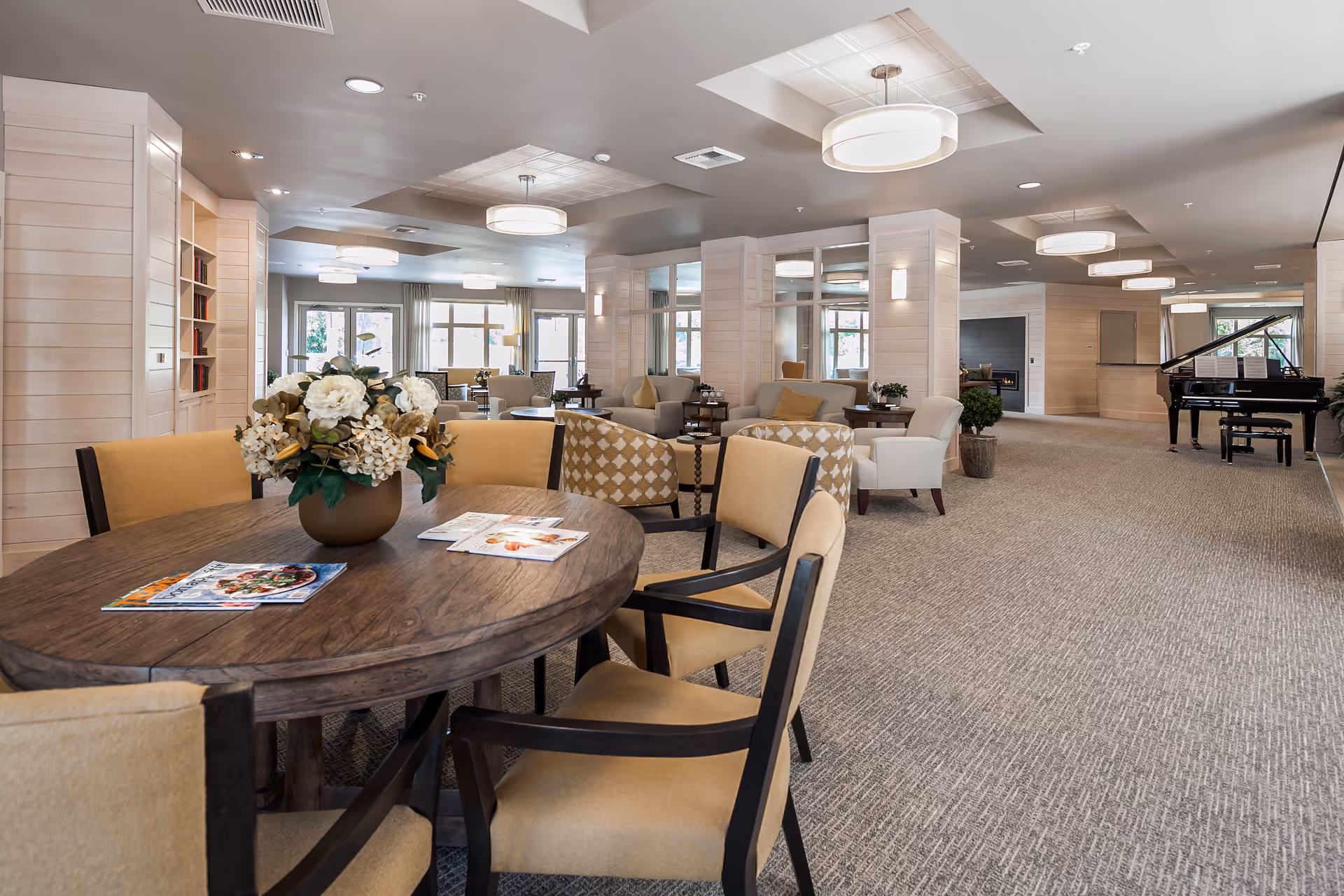 A spacious and well-lit senior living facility common area with a round wooden table and beige cushioned chairs in the foreground. The table has a floral centerpiece and magazines on it. The room features multiple seating areas with armchairs and small tables, large windows letting in natural light, modern ceiling lights, and a grand piano in the background.