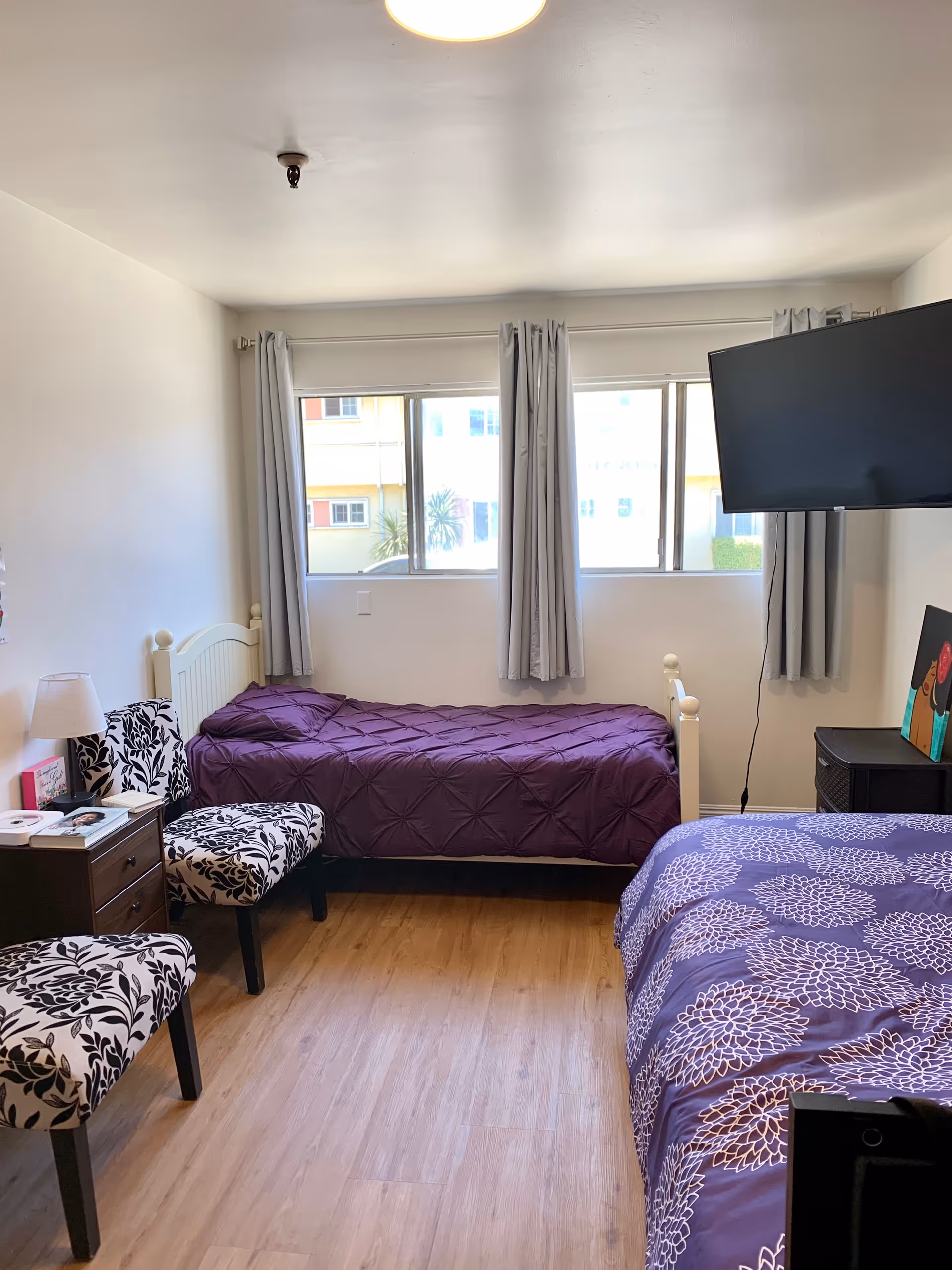 A bright room with two single beds covered in purple bedspreads, a wooden floor, two patterned chairs, a small wooden nightstand with a lamp and books, a wall-mounted flat screen TV, and a window with light gray curtains letting in natural light.