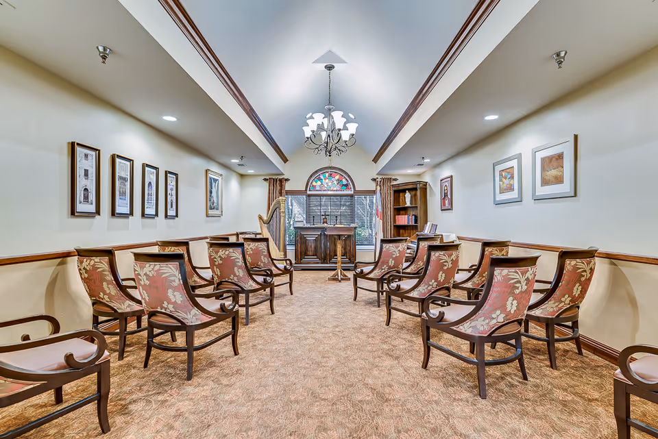 A small chapel or meeting room with two rows of wooden chairs with floral upholstery facing a wooden podium and a harp. The room has beige walls adorned with framed pictures, a chandelier hanging from the ceiling, and a stained glass window behind the podium.