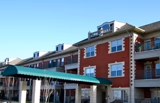 Exterior view of a multi-story senior living community building with red brick and beige stone accents, featuring balconies and a green canopy entrance.