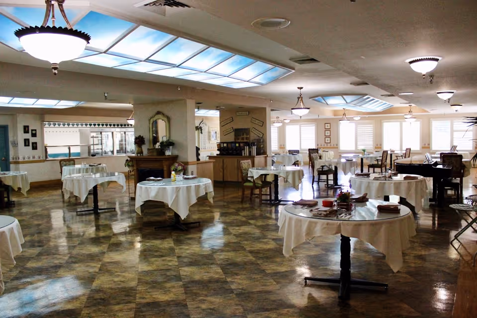 A spacious dining room with multiple round tables covered with white tablecloths, each set with napkins and some with small centerpieces. The room has a polished floor, large windows with blinds, ceiling lights, and a skylight. There is a piano and some chairs along the walls, and a coffee station is visible in the background.
