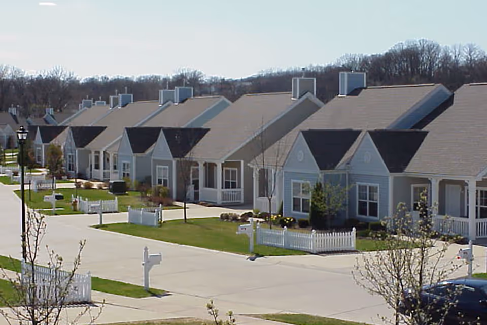 A row of single-story residential buildings with pitched roofs and small front porches, each surrounded by white picket fences and small lawns. The street in front is paved and there are some young trees planted along the sidewalks. The background shows leafless trees under a clear sky.