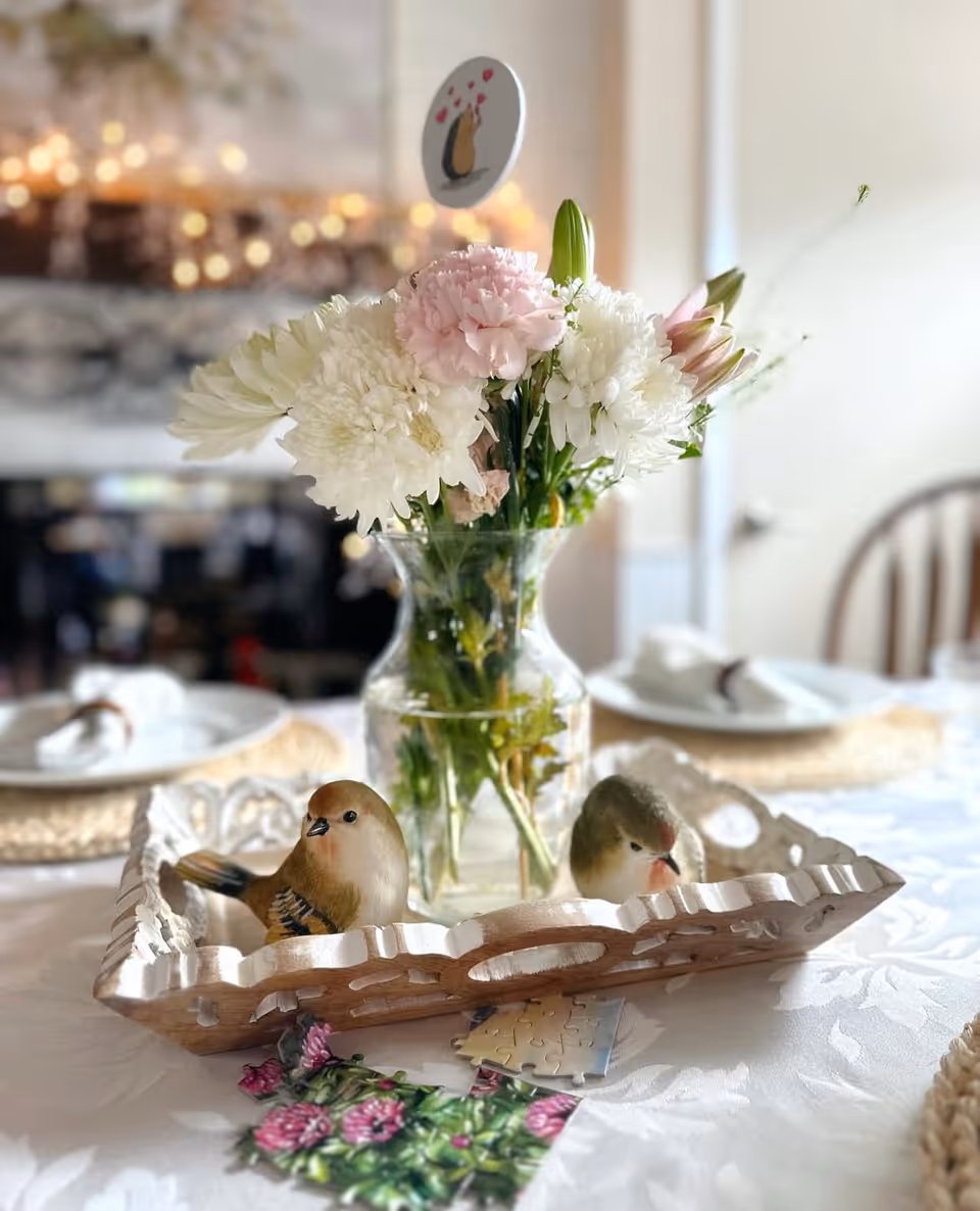 A close-up of a dining table centerpiece featuring a glass vase with white and pink flowers, placed inside a decorative wooden tray with two small bird figurines. The table is set with woven placemats, white plates, and napkins. The background shows a softly lit room with blurred lights and a wooden chair.