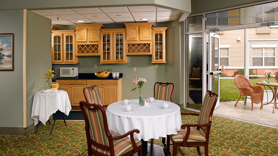 A cozy dining area with a round table covered with a white tablecloth and four wooden chairs with striped upholstery. The table is set with cups, saucers, and a small vase of flowers. Behind the table is a kitchenette with wooden cabinets, a microwave, and a fruit bowl. To the right, a glass door opens to an outdoor patio with wicker chairs and a small table. The room has green walls and patterned carpet.