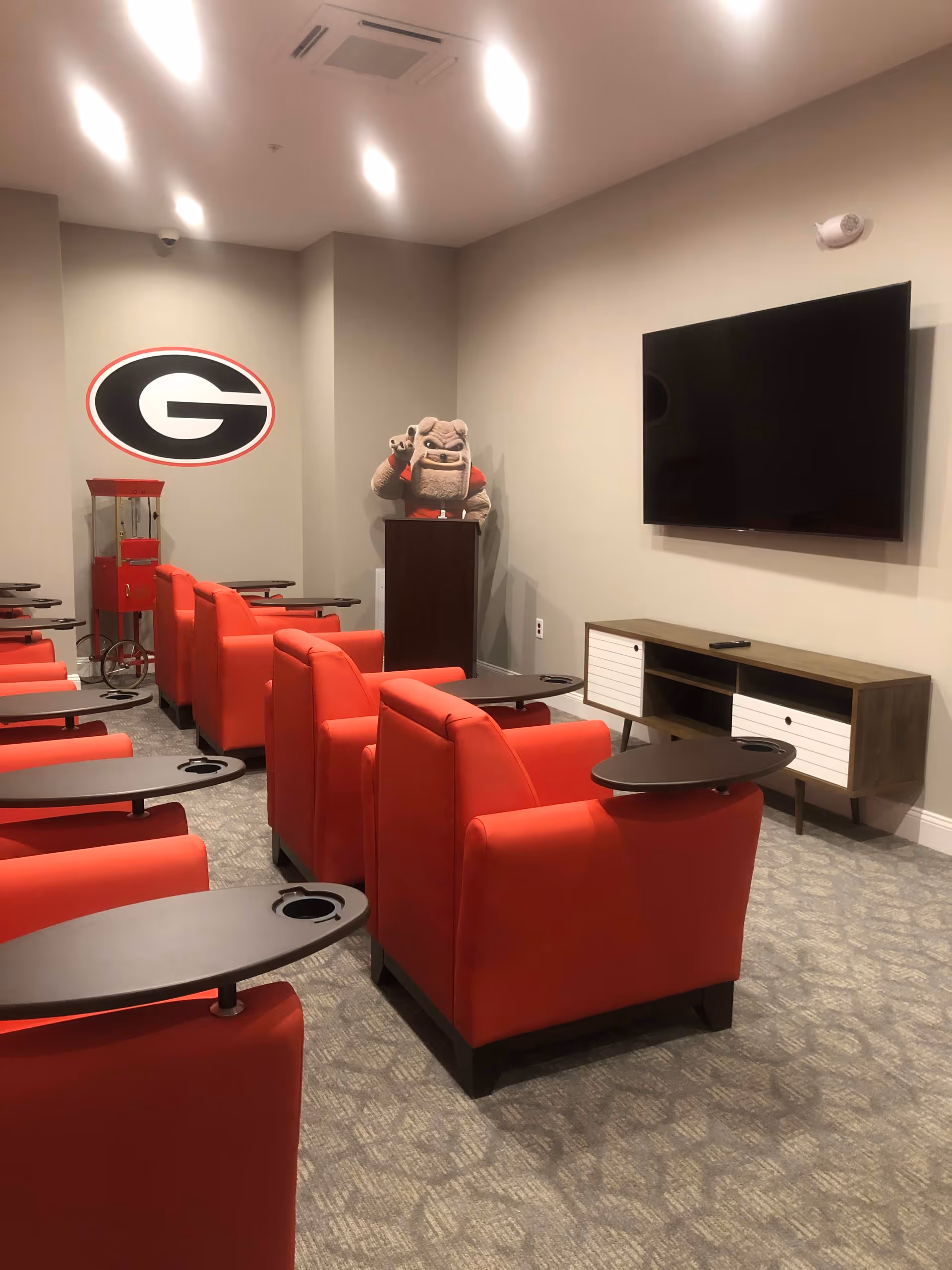Media room with rows of red reclining chairs facing a wall-mounted TV, a popcorn cart, and a bulldog mascot beside a large G logo on the wall.