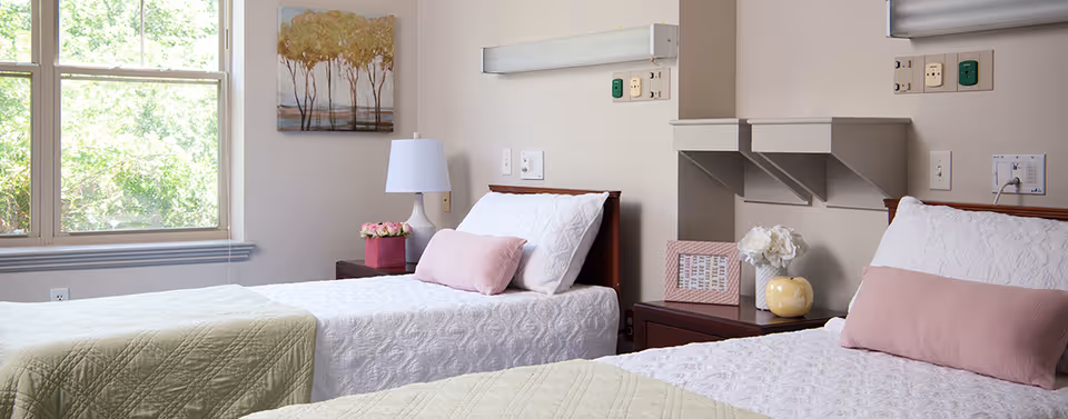 Two neatly made twin beds with pastel pillows, bedside tables, a lamp, and a window in a bright, softly decorated senior living bedroom.