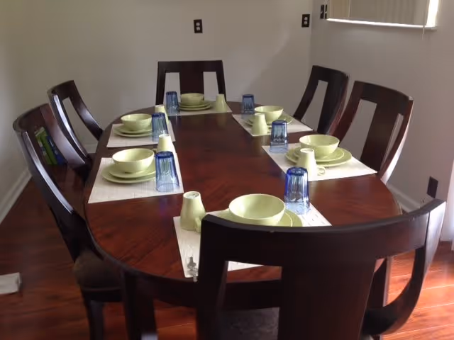 A dark wood dining table in a small dining room arranged with light green dishes, blue glasses, and placemats and surrounded by wooden chairs.