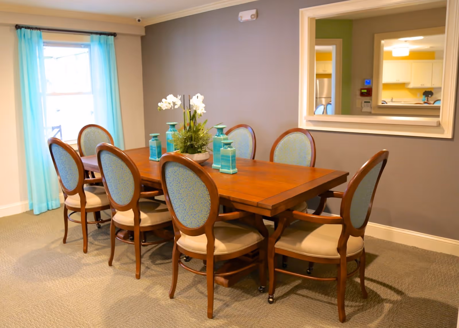 A dining room with a wooden table surrounded by eight upholstered chairs with light blue patterned backs and beige seats. The table is decorated with a floral centerpiece and four turquoise decorative jars. A large window with sheer turquoise curtains is on the left wall, and a large mirror hangs on the right wall reflecting a kitchen area.