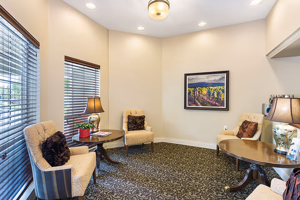 Cozy sitting area with upholstered chairs, side tables, lamps and framed art beside large windows in a retirement community common room.