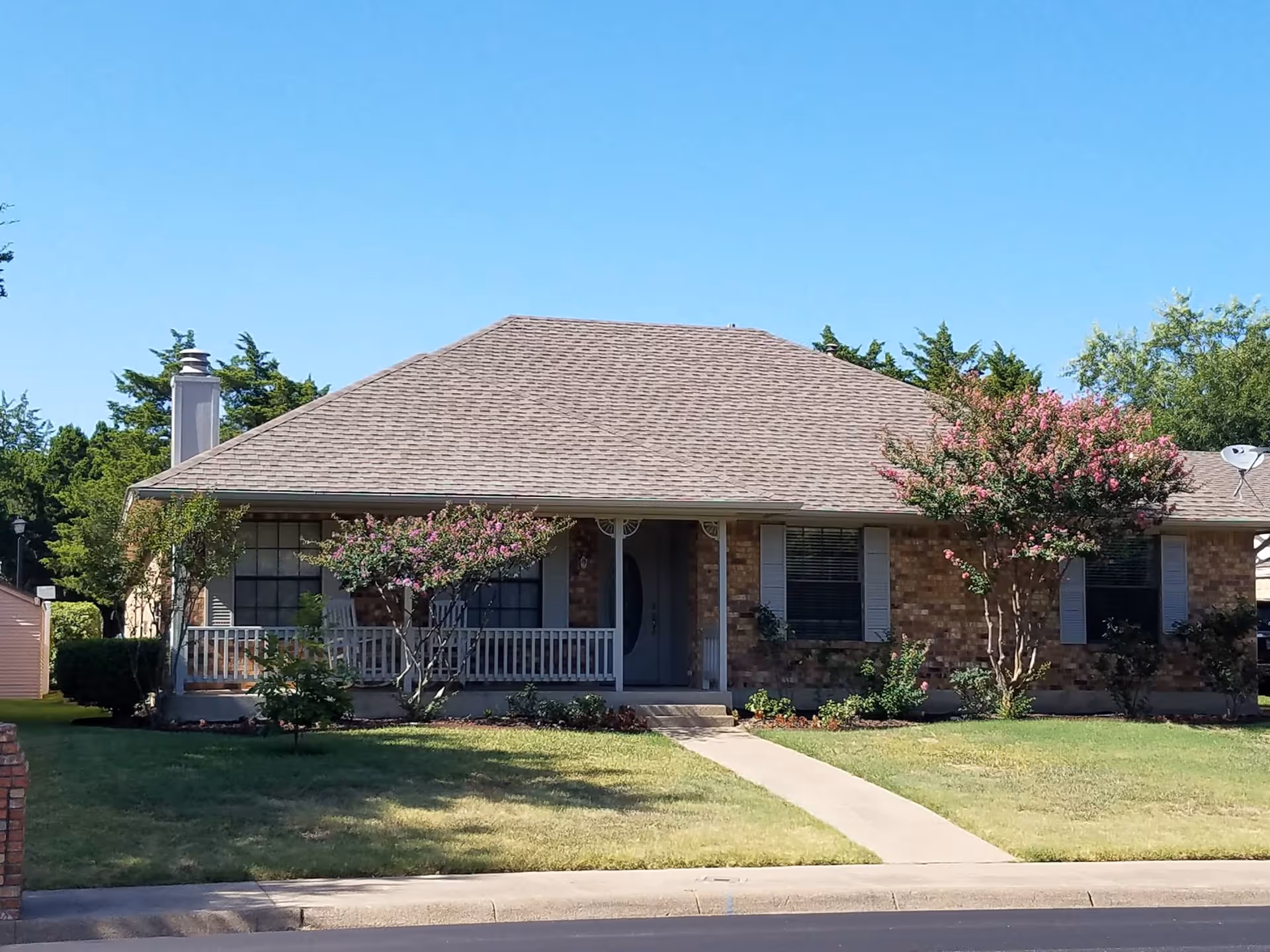 Single-story brick house with a covered front porch, walkway, lawn, and flowering trees under a clear blue sky.