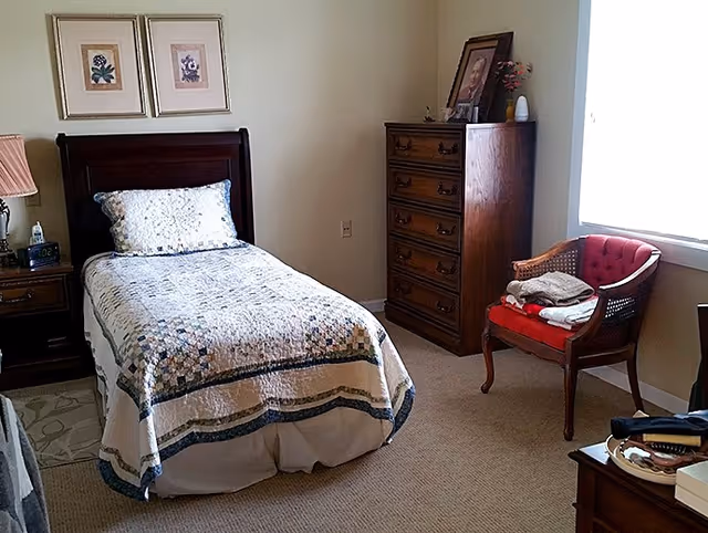 A small bedroom with a single bed covered with a patterned quilt. Next to the bed is a wooden nightstand with a lamp, clock, and hand sanitizer. On the wall above the bed are two framed floral pictures. Across from the bed is a tall wooden dresser with a framed photo and decorative items on top. A red cushioned wooden chair with folded clothes sits near a window with white blinds, letting in natural light. The room has beige carpet and light-colored walls.