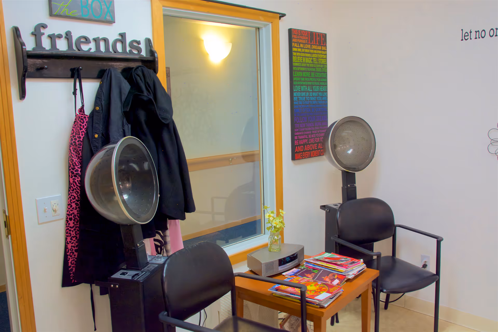 Small salon-style interior with two hooded hair dryers, two black chairs, a small table piled with magazines, coats hanging on a wall rack and decorative signs.