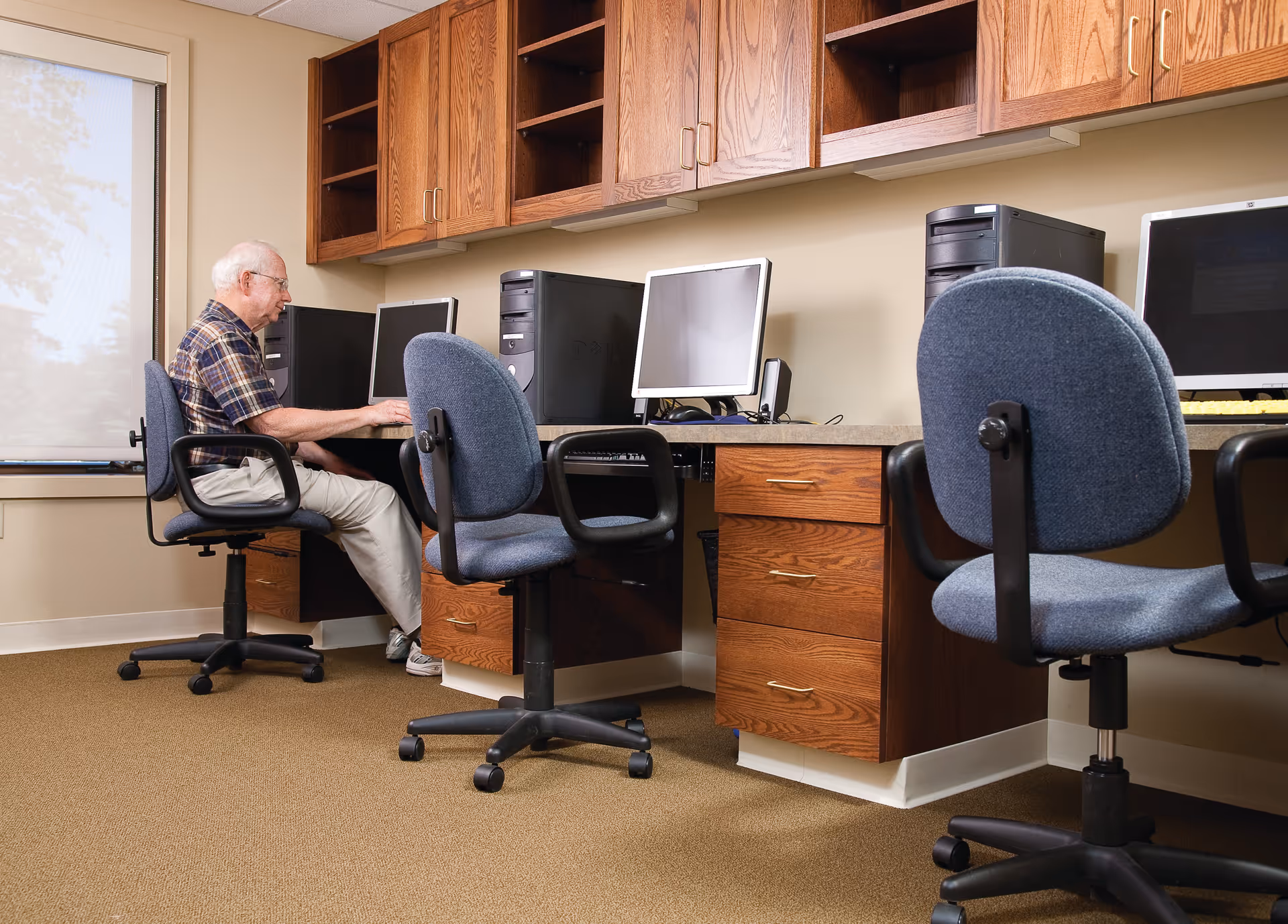 An elderly man sits at a desk in a communal computer room using a desktop computer, with several empty workstations and blue swivel chairs.