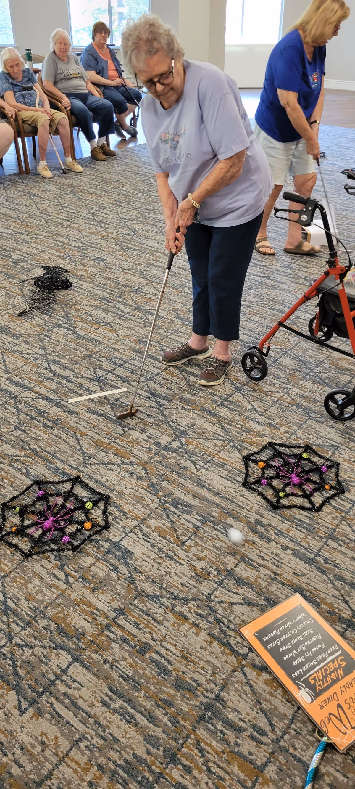 An elderly woman playing indoor mini golf in a senior living facility common area, with other seated elderly individuals watching in the background. The floor is carpeted and decorated with Halloween-themed spider web targets. Another elderly woman with a walker is also visible nearby.