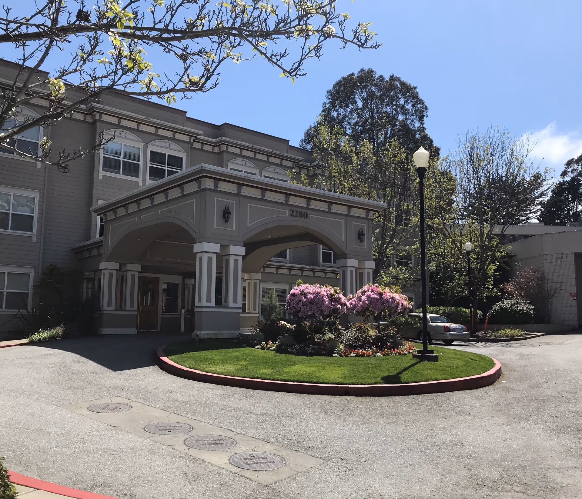 Exterior view of a senior living facility entrance with a covered drop-off area, manicured lawn, blooming pink flowers, trees, and a clear blue sky.