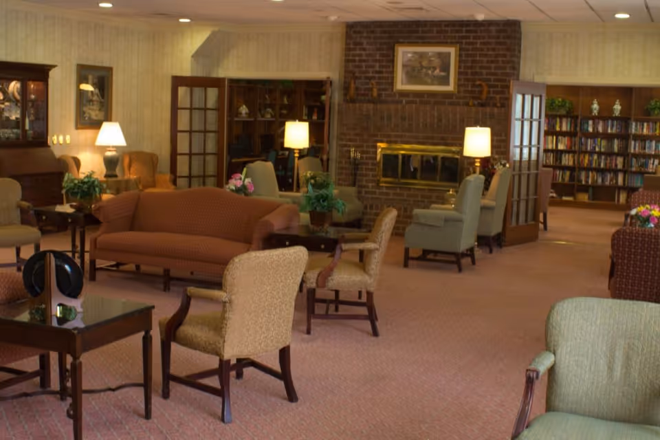 A cozy senior living facility common area with multiple upholstered chairs and sofas arranged around small tables. The room features a brick fireplace with a framed picture above it, several table lamps providing warm lighting, and bookshelves filled with books in the background. The space has a carpeted floor and wallpapered walls, creating a comfortable and inviting atmosphere.