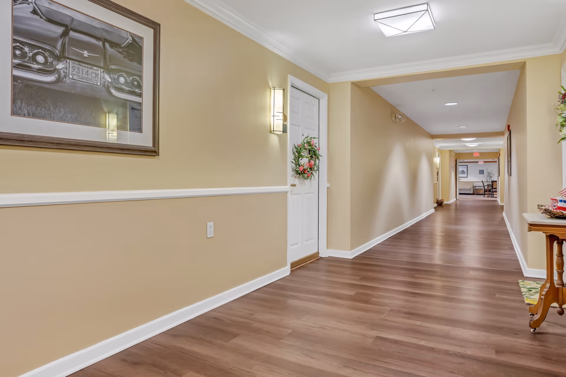 A long, clean hallway with light beige walls and wooden flooring. There is a white door decorated with a floral wreath on the left side, a framed black and white photo of a vintage car above a wall light, and a small wooden table with a floral arrangement on the right. The hallway is well-lit with ceiling lights and wall sconces, leading to a seating area in the distance.