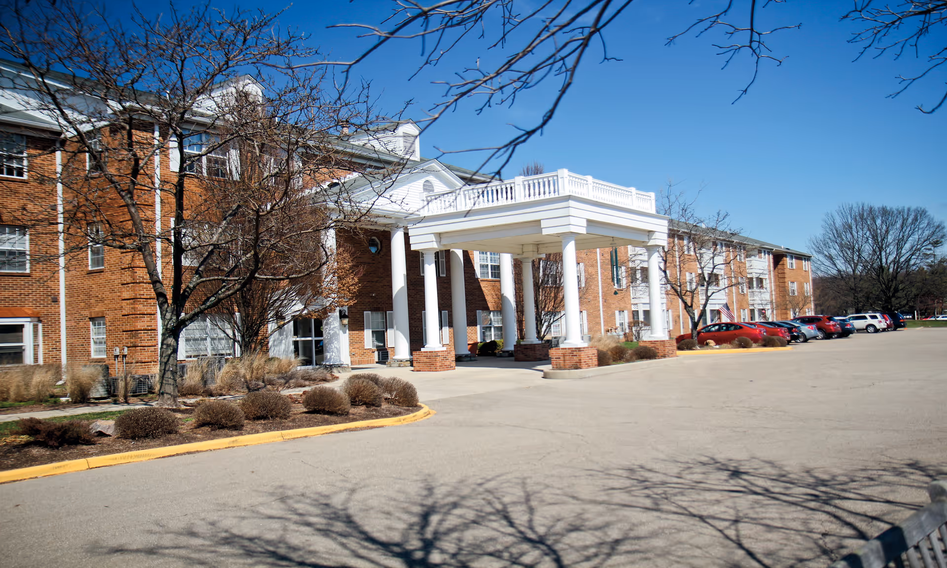 Exterior view of a large brick senior living facility with a white columned entrance canopy, several leafless trees, and a parking lot with multiple cars under a clear blue sky.