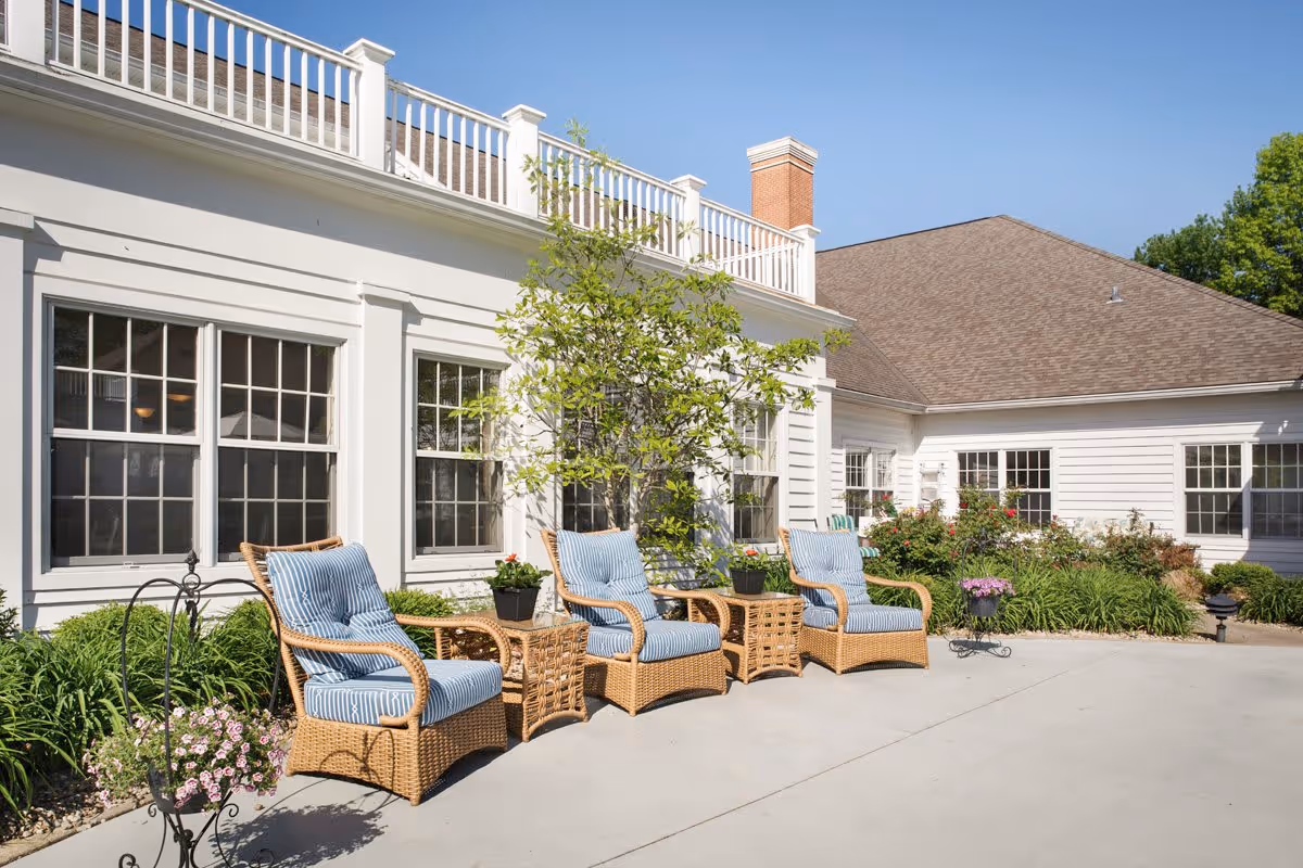 Outdoor patio area at StoryPoint Gahanna Central featuring three wicker chairs with blue striped cushions and two wicker side tables. The patio is adjacent to a white building with multiple windows, surrounded by green plants and flowers under a clear blue sky.