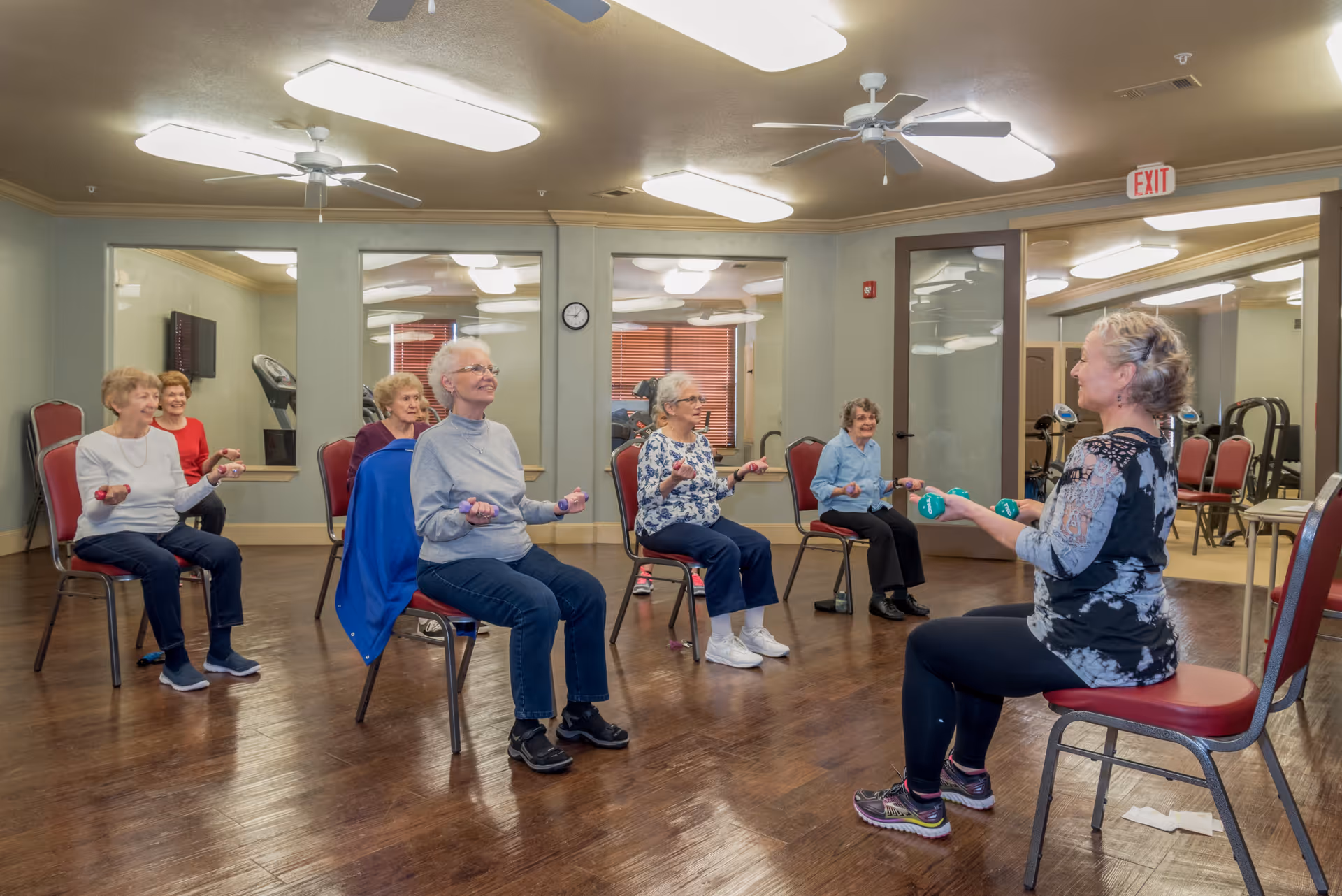 A group of elderly women seated on chairs in a fitness room participating in a seated exercise class using small hand weights, led by an instructor also seated and holding weights. The room has wooden floors, large mirrors on the walls, ceiling fans, and bright overhead lighting.
