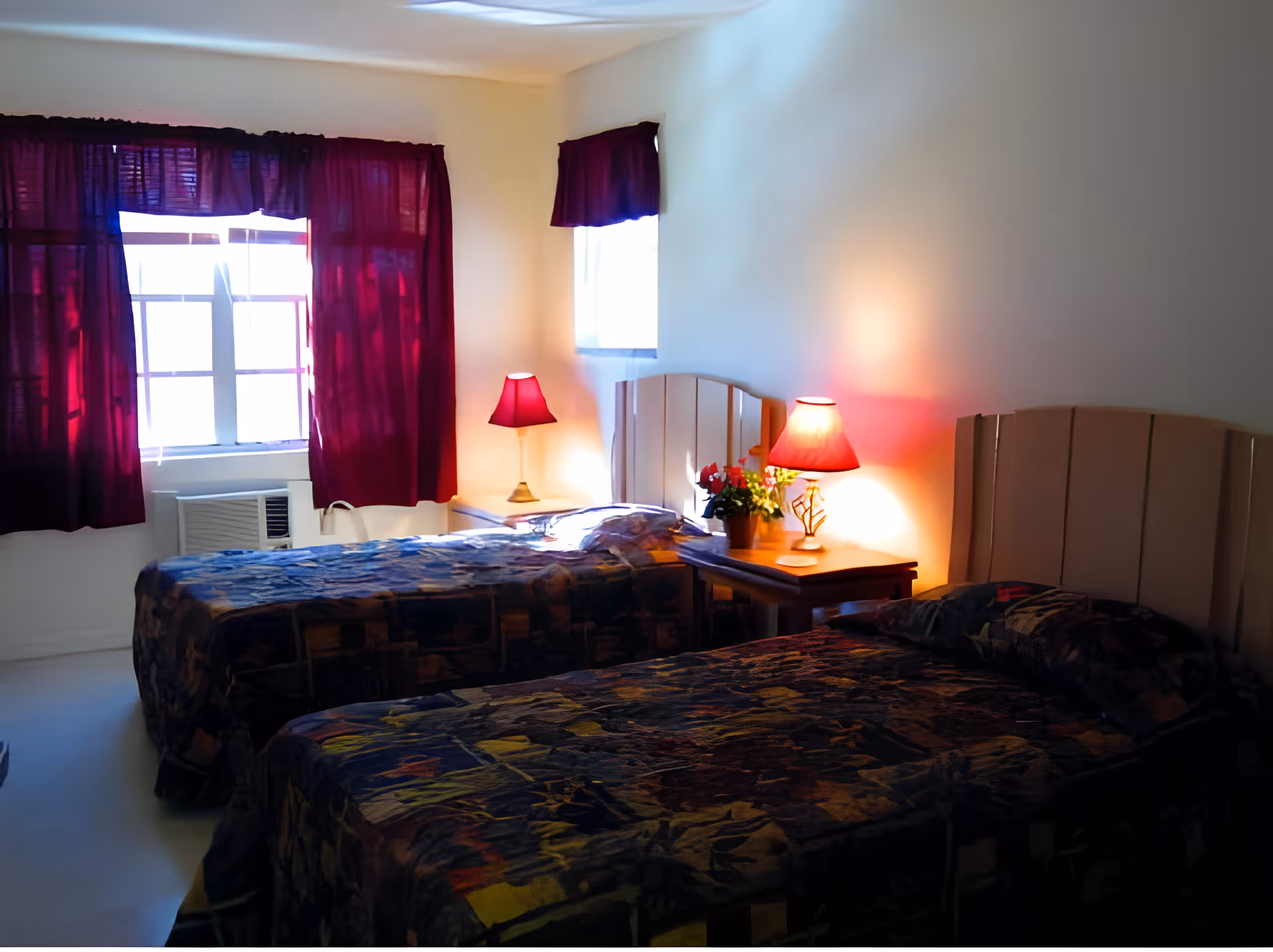 A bedroom with two twin beds covered in dark patterned bedspreads. Each bed has a light-colored headboard. Between the beds is a wooden nightstand with a red lampshade and a small potted plant. The room has two windows with dark red curtains, and an air conditioning unit is installed below one window.